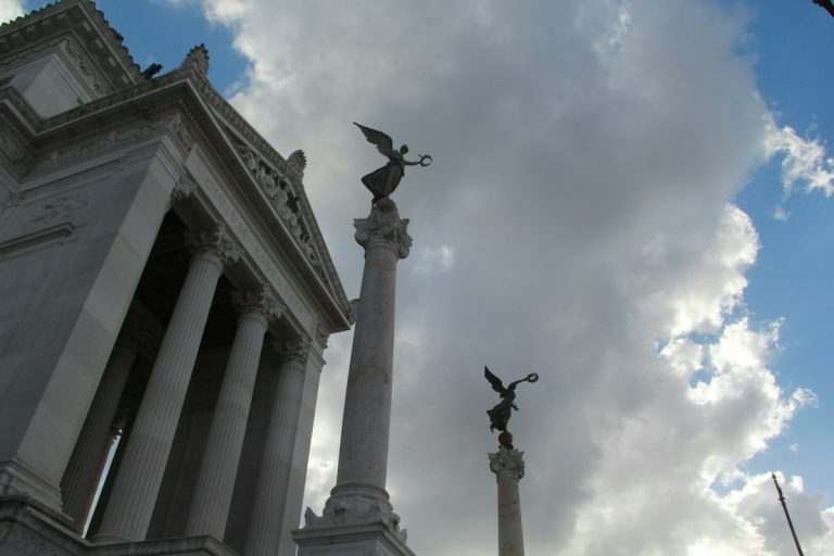 Two winged statues atop columns against cloudy sky